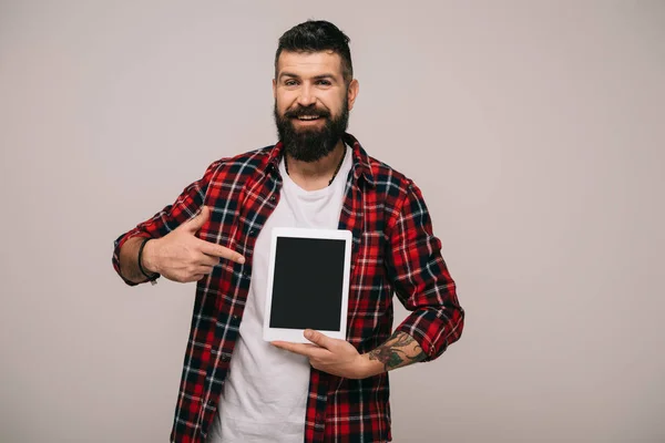 Улыбающийся бородатый мужчина указывает на цифровой планшет с чистым экраном, изолированный на сером — стоковое фото