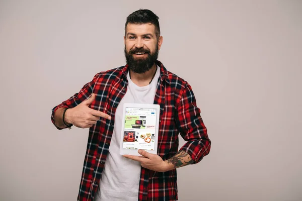 Sonriente hombre guapo con camisa a cuadros apuntando a la tableta digital con aplicación ebay, aislado en gris - foto de stock