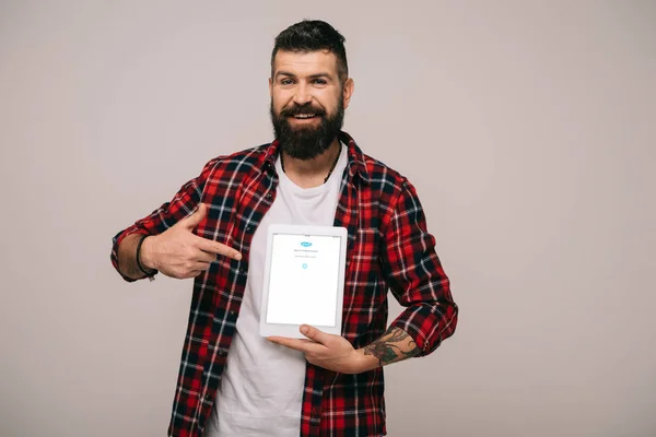 Bärtiger Mann im karierten Hemd zeigt auf digitales Tablet mit Skype-App, isoliert auf grau — Stockfoto