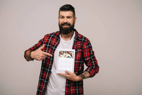Бородатый мужчина в клетчатой рубашке, указывая на цифровой планшет с четырехквадратным приложением, изолированным на сером — стоковое фото