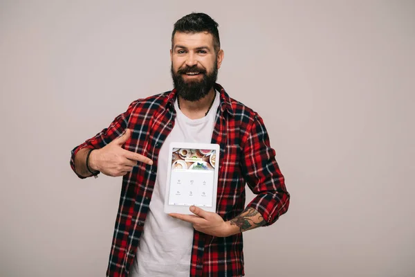 Улыбающийся мужчина в клетчатой рубашке указывает на цифровой планшет с четырехквадратным приложением, изолированным на сером — стоковое фото