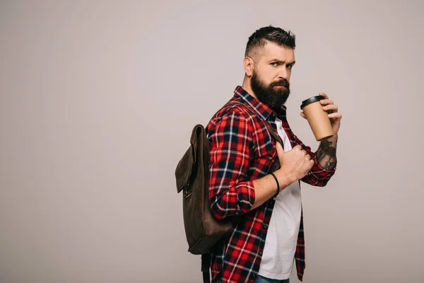 Homme barbu sérieux en chemise à carreaux avec sac à dos tenant café pour aller isolé sur gris — Photo de stock