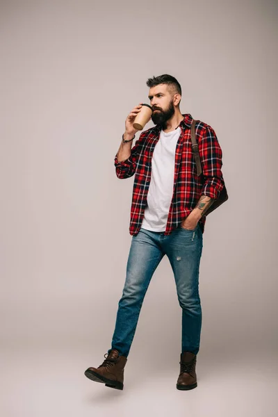 Uomo barbuto in camicia a scacchi che cammina e beve caffè per andare isolato sul grigio — Foto stock