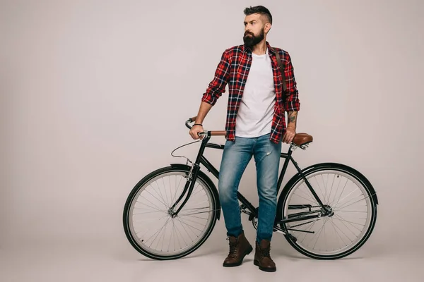 Bearded man in checkered shirt standing at bike on grey — Stock Photo