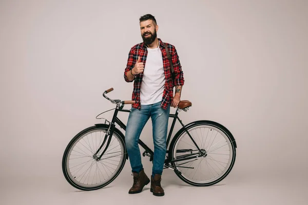 Guapo hombre sonriente con camisa a cuadros posando con bicicleta y mostrando el pulgar hacia arriba en gris - foto de stock