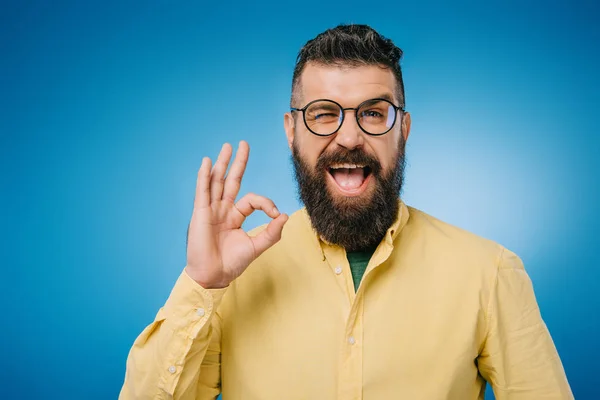 Щасливий бородатий чоловік в окулярах підморгує і показує знак ОК, ізольований на синьому — стокове фото