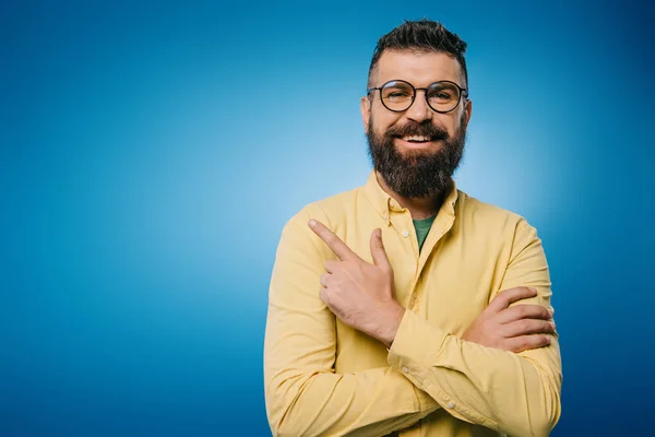 Hombre barbudo alegre en gafas apuntando aislado en azul - foto de stock