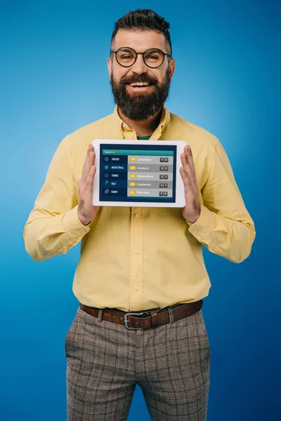 Улыбающийся бородатый мужчина показывает цифровой планшет со спортивным приложением, изолированным на синий — стоковое фото