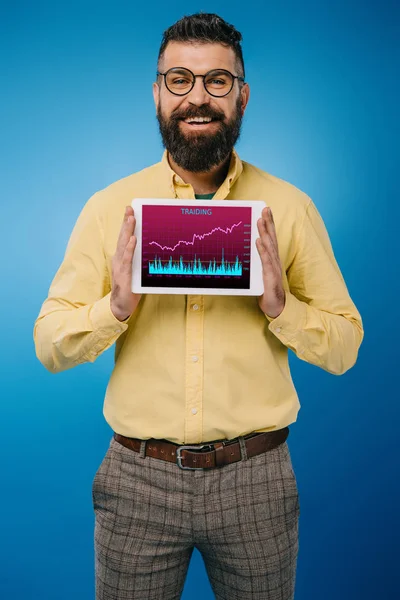Sonriente hombre barbudo presentando tableta digital con aplicación tariding, aislado en azul - foto de stock