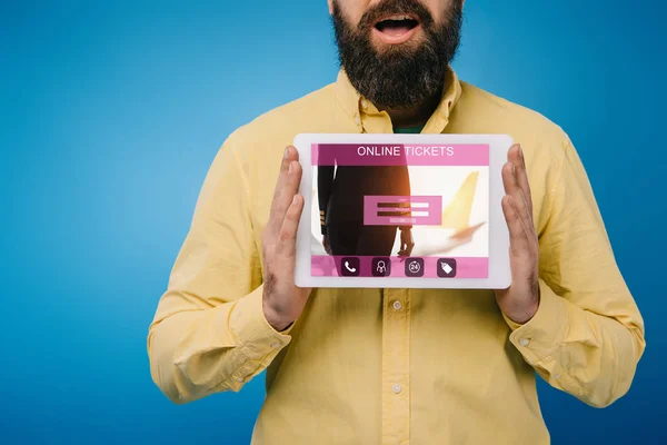 Vista recortada del hombre barbudo conmocionado mostrando tableta digital con aplicación de boletos en línea, aislado en azul - foto de stock