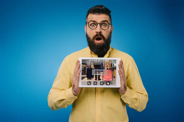 Sorprendido barbudo hombre que presenta tableta digital con aplicación de reserva en línea, aislado en azul - foto de stock