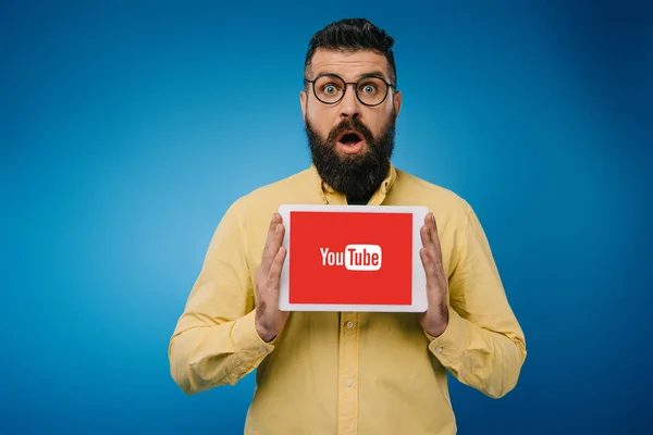 Homem barbudo surpreso mostrando tablet digital com aplicativo youtube, isolado em azul — Fotografia de Stock