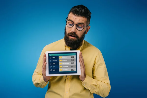 Hombre barbudo interesado mirando tableta digital con aplicación deportiva, aislado en azul - foto de stock