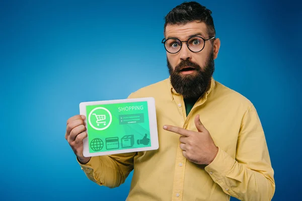 Шокированный бородатый мужчина указывает на цифровой планшет с помощью торгового приложения, изолированного на синий — стоковое фото