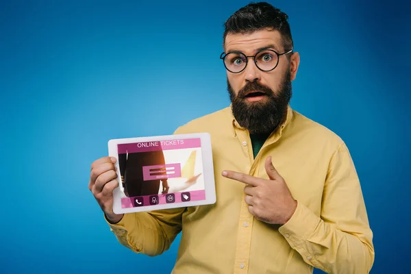 Шокированный бородатый мужчина указывает на цифровой планшет с приложением онлайн-билетов, изолированный на синий — стоковое фото