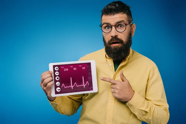 Sorprendido hombre barbudo apuntando a la tableta digital con aplicación de datos de salud, aislado en azul - foto de stock