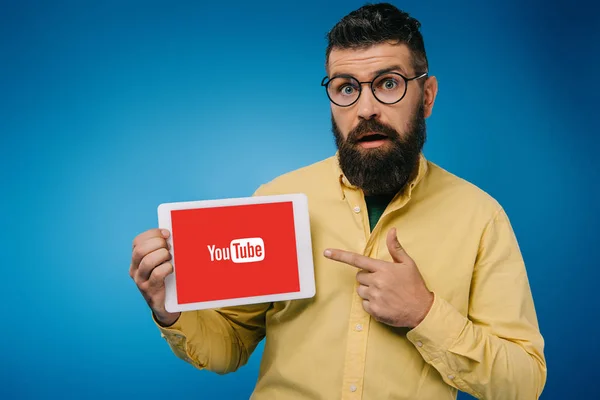 Hombre barbudo sorprendido apuntando a la tableta digital con aplicación de youtube, aislado en azul - foto de stock