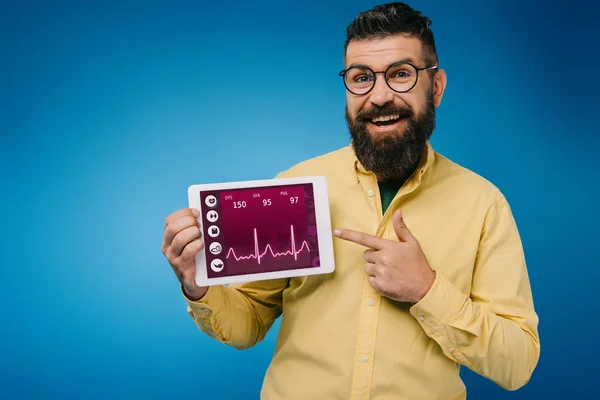 Felice uomo barbuto che punta al tablet digitale con i dati di salute, isolato su blu — Foto stock