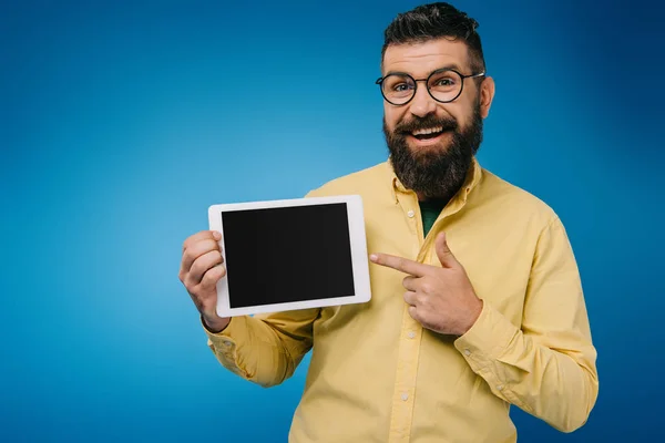 Красивый улыбающийся мужчина указывает на цифровой планшет с чистым экраном, изолированный на синий — стоковое фото