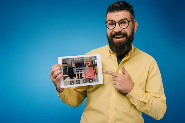 Glücklicher bärtiger Mann zeigt auf digitales Tablet mit Online-Buchungs-App, isoliert auf blauem Grund — Stockfoto