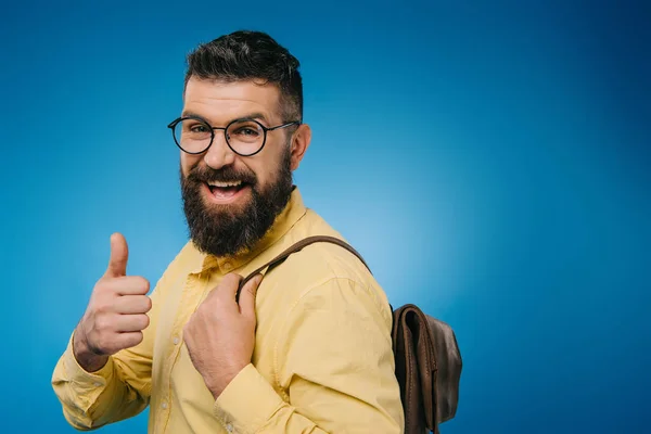 Веселий бородатий чоловік з рюкзаком, що показує великий палець вгору, ізольований на синьому — стокове фото