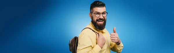 Smiling bearded man with backpack showing thumb up, isolated on blue — Stock Photo