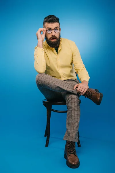 Surpris bel homme aux lunettes assis sur une chaise isolée sur bleu — Photo de stock