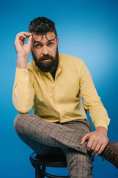 Бородатий чоловік в окулярах сидить на стільці ізольовано на синьому — стокове фото