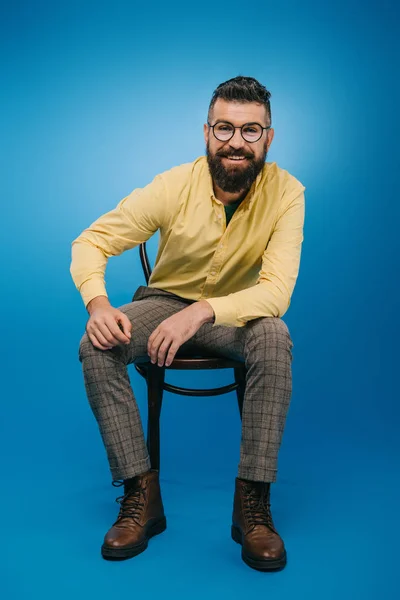 Homme barbu souriant dans des lunettes assis sur une chaise isolée sur bleu — Photo de stock