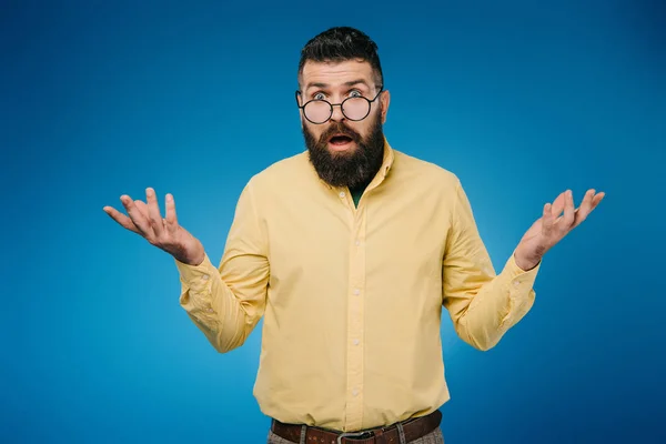 Homme barbu dans des lunettes avec geste haussant les épaules isolé sur bleu — Photo de stock