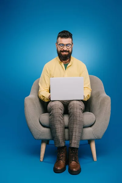 Happy bearded businessman sitting in armchair and using laptop on blue — Stock Photo