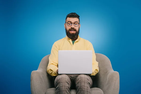 Uomo d'affari eccitato utilizzando il computer portatile mentre seduto in poltrona, isolato sul blu — Foto stock