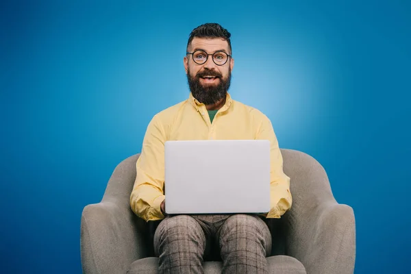 Щасливий бородатий бізнесмен, використовуючи ноутбук, сидячи в кріслі, ізольований на синьому — стокове фото