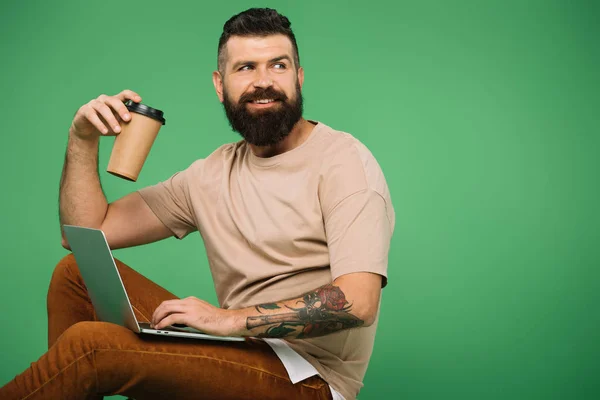 Happy bearded man with coffee to go using laptop isolated on green — Stock Photo