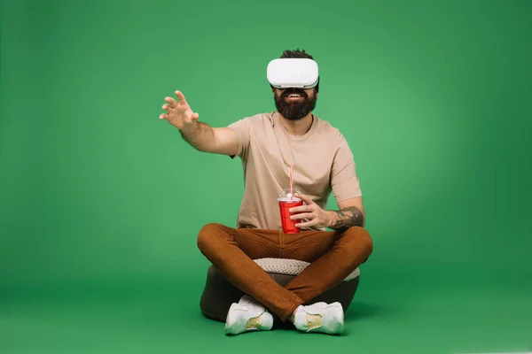 Бородатий чоловік з паперовим склом содової з використанням гарнітури віртуальної реальності ізольовано на зеленому — стокове фото