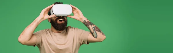 Hombre barbudo sonriente usando auriculares de realidad virtual aislados en verde - foto de stock