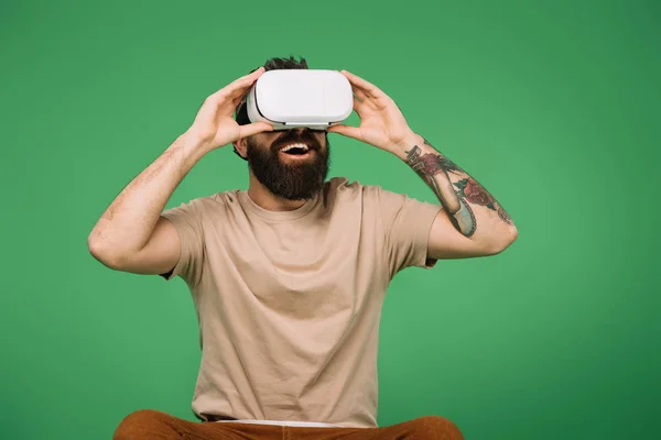 Hombre barbudo excitado usando auriculares de realidad virtual aislados en verde - foto de stock