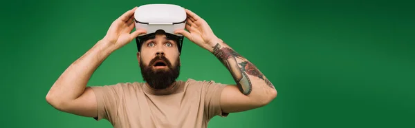 Uomo barbuto scioccato con auricolare realtà virtuale isolato su verde — Foto stock