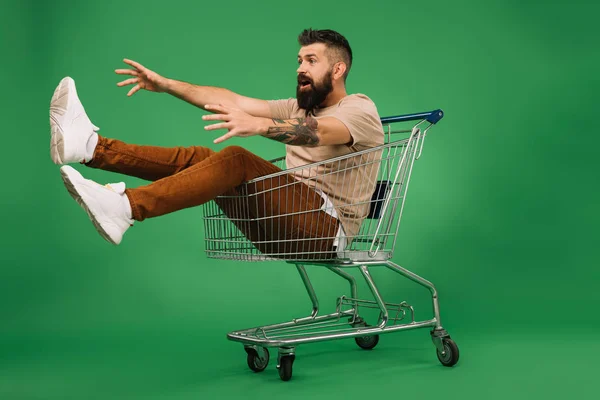 Excited man gesturing and sitting in shopping cart isolated on green — Stock Photo