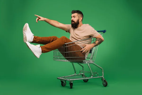 Aufgeregter gutaussehender Mann zeigt etwas, während er im Einkaufswagen isoliert auf Grün sitzt — Stockfoto