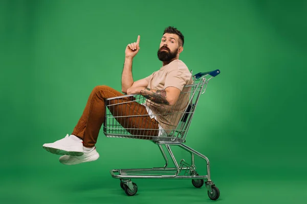 Divertente uomo barbuto che punta verso l'alto e seduto nel carrello della spesa isolato su verde — Foto stock