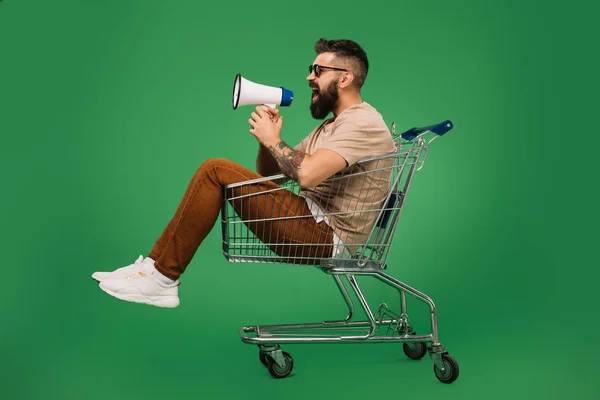 Bearded man shouting into megaphone while sitting in shopping cart isolated on green — Stock Photo