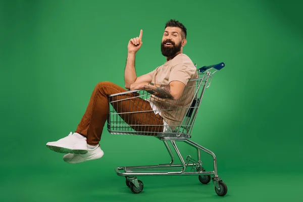 Sonriente hombre apuntando hacia arriba y sentado en el carro de la compra aislado en verde - foto de stock