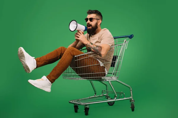 Bearded man with megaphone sitting in shopping cart isolated on green — Stock Photo
