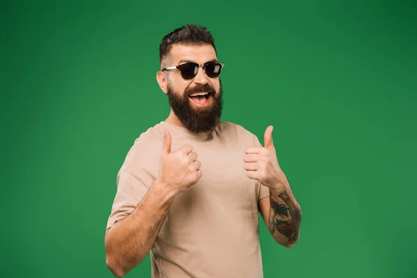 Bearded man in sunglasses showing thumbs up, isolated on green — Stock Photo