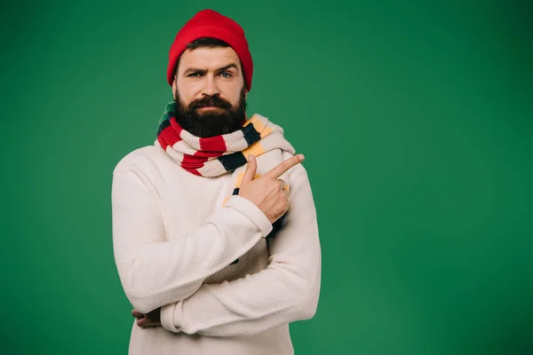 Sick bearded man in hat and scarf pointing aside isolated on green — Stock Photo