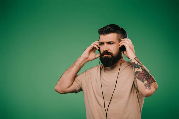 Hombre tatuado barbudo escuchando música en auriculares aislados en verde - foto de stock