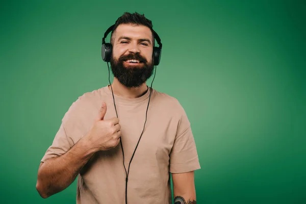 Bearded smiling man showing thumb up while listening music in headphones isolated on green — Stock Photo