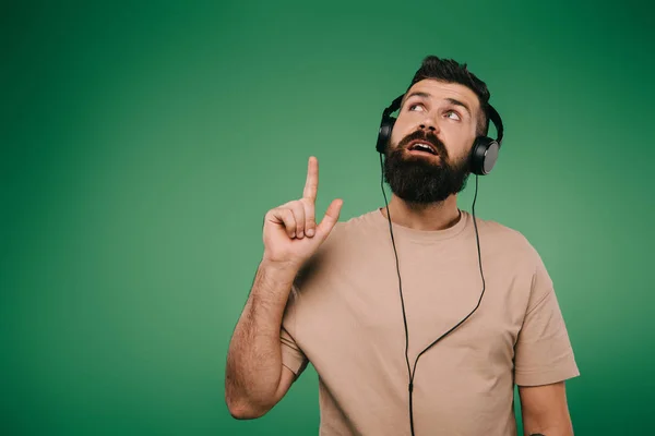 Bärtiger Mann mit Kopfhörer, der eine Idee hat und nach oben zeigt, isoliert auf grün — Stockfoto