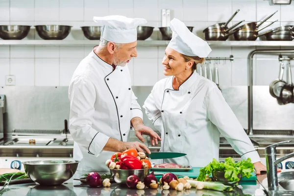 Female and male chefs in uniform and hats looking at each other during cooking in restaurant kitchen — Stock Photo